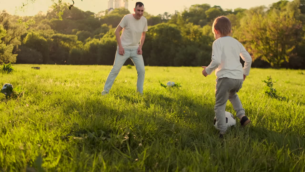 A young father in a white t-shirt with two sons playing football on the grass at sunset in the sun in slow motion