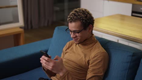 cheerful young man web surfing on his mobile phone on the sofa at home