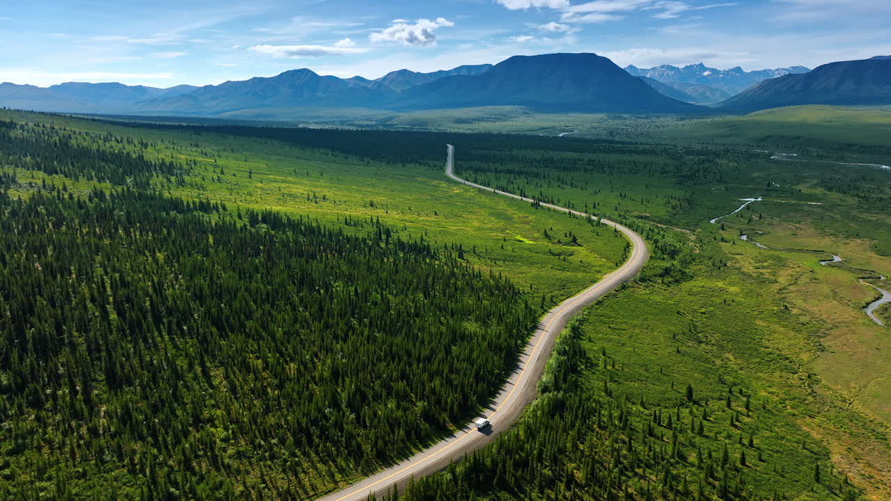 Campervan rides by the wavy road crossing the green valley. Spectacular mountain range at backdrop. Alaska wilderness. Aerial view