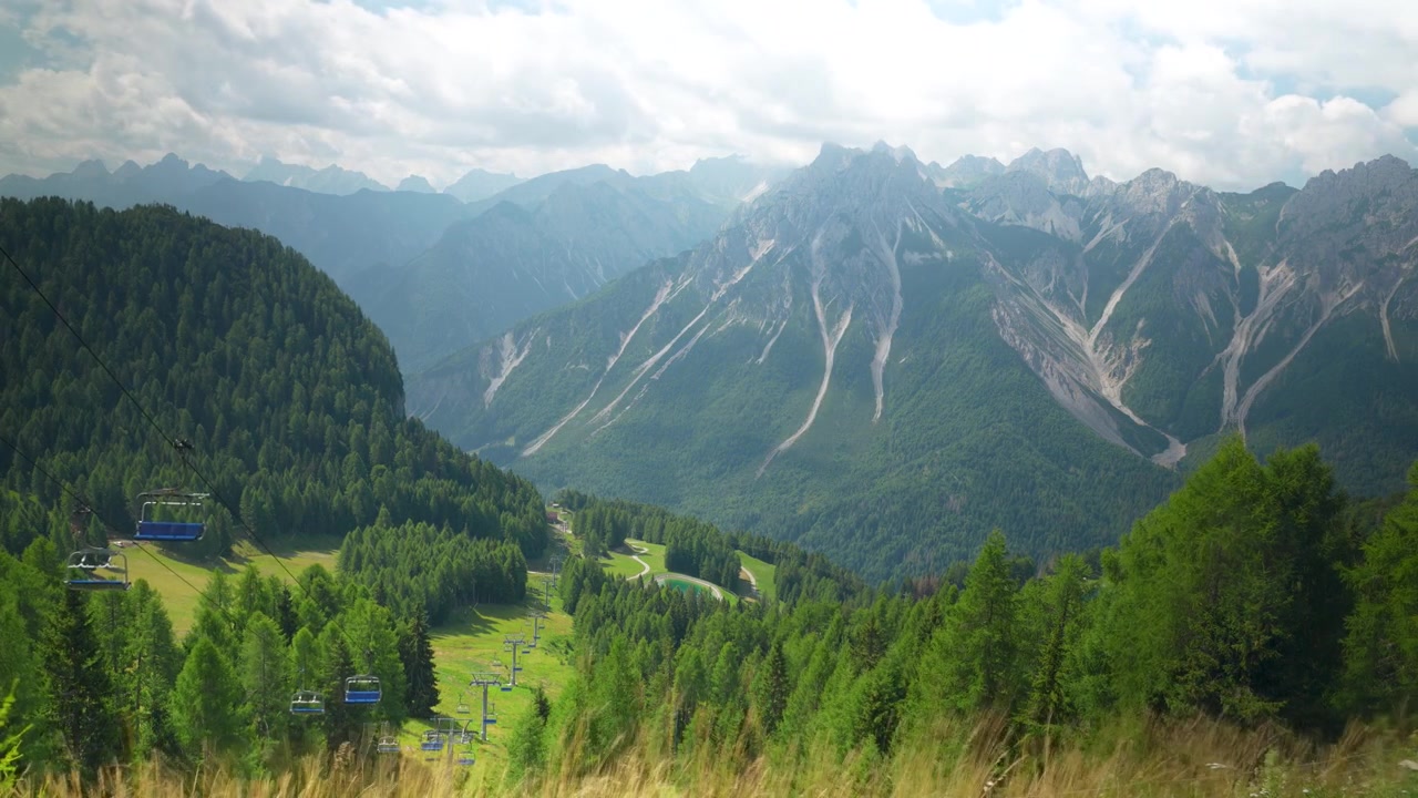 Timelapse overlooking Malga Varmost with lush alpine forests, winding chairlift, and dramatic Dolomite mountains beneath fast-moving clouds in northern Italy