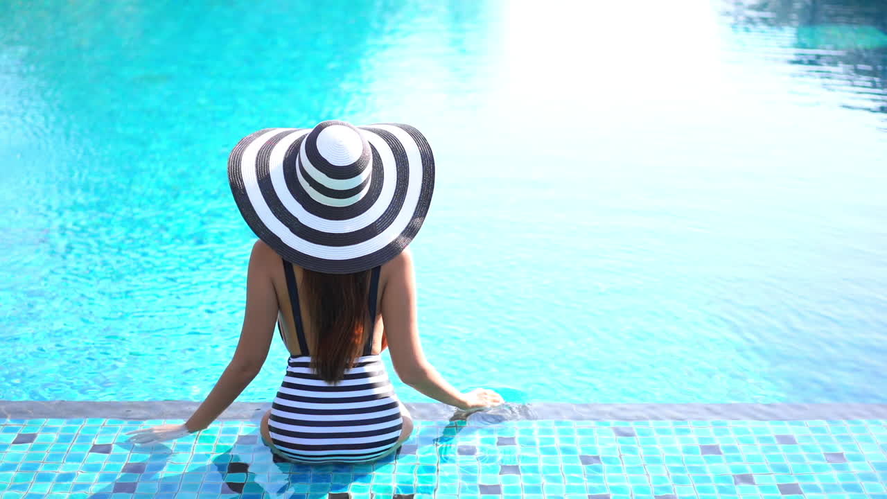Back view of slim sexy girl wearing black and white striped swimming suit and sun hat sitting on the edge of the swimming pool, top view slow-motion