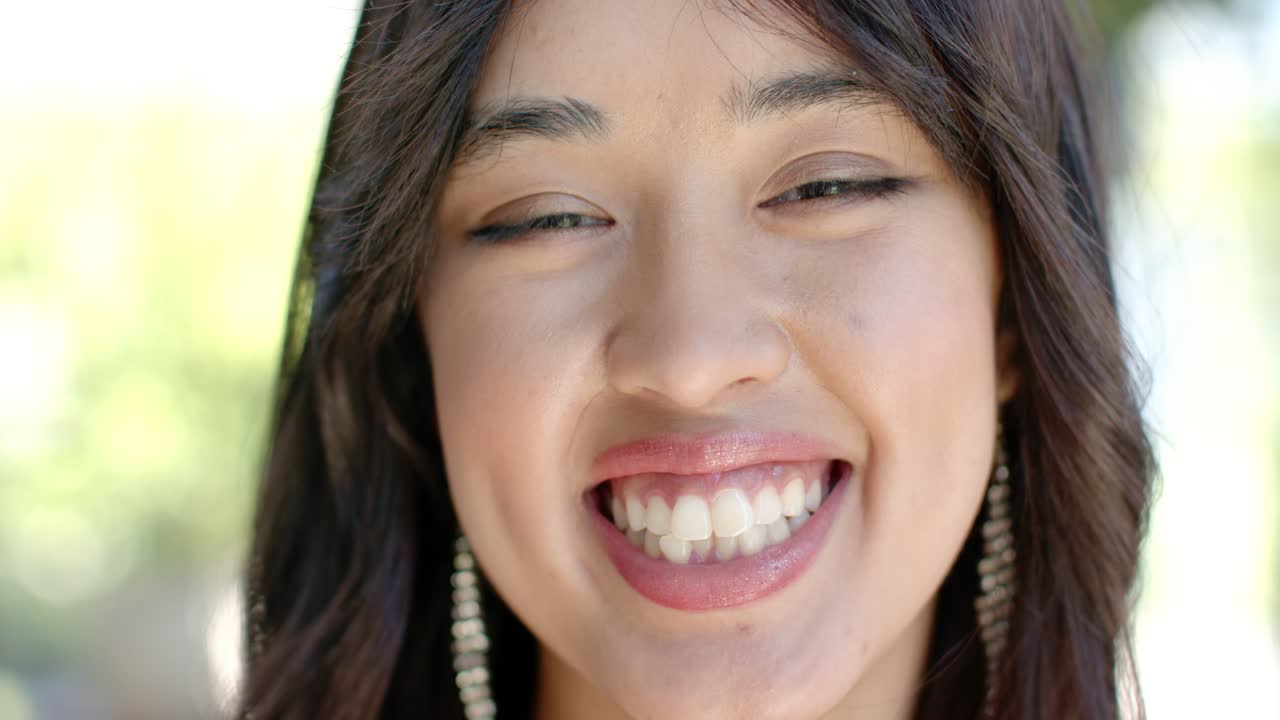 Woman 20s responding to photographer prompt, smiling, squinting, showing earrings outdoors