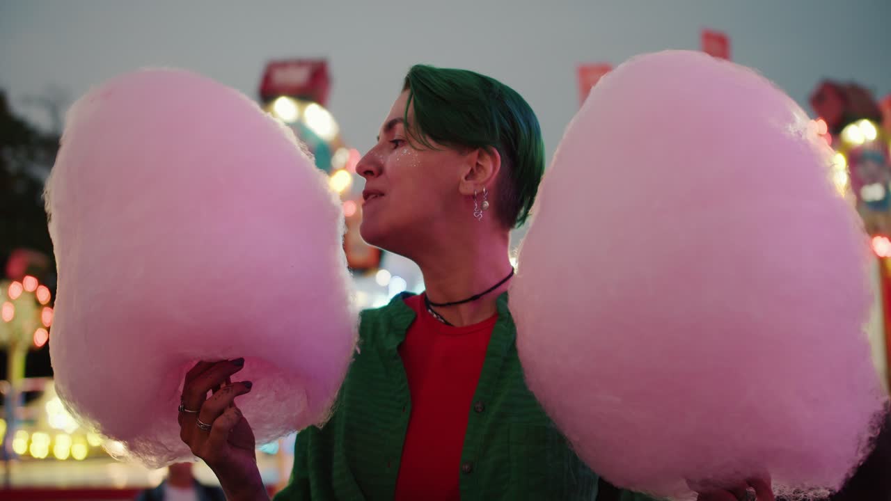 초록색 셔츠를 입고 짧은 초록색 헤어스타일을 가진 레즈비언 소녀는 그녀의 손에 거대한 분홍색 면화 사탕을 들고 밝은 놀이 공원에서 산책하는 동안 그것을 습니다.