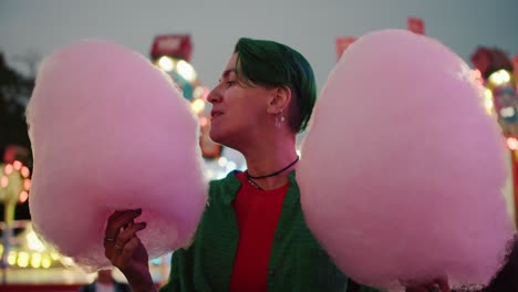 A-lesbian-girl-with-a-short-green-haircut-in-a-Green-shirt-holds-huge-pink-cotton-candy-in-her-hands-and-bites-into-it-during-her-walk-in-a-bright-amusement-park