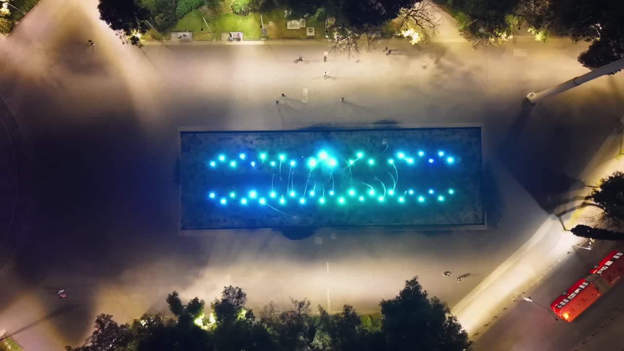 Plano de estabelecimento de drone de uma fonte na praça com luzes coloridas à noite.