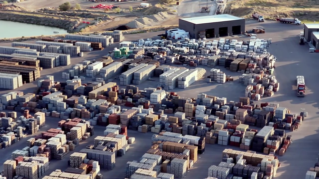 Stacks of masonry blocks, segmental retaining walls, pavers, and fence blocks at a busy outdoor facility under warm, golden sunlight. No people visible. Aerial parallax.