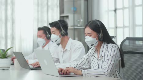 three asian call center agents wearing headsets and masks sitting on the chairs and typing on computers while speaking to customers on the call at the office