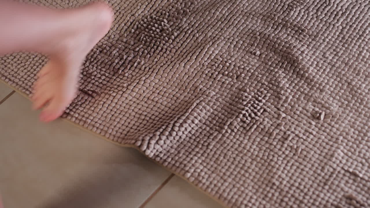 High angle view shows female feet stepping onto soft bath mat near bathtub, toes pressing plush texture on tiled floor, water drops visible, slip prevention, cozy spa vibe in clean home bathroom