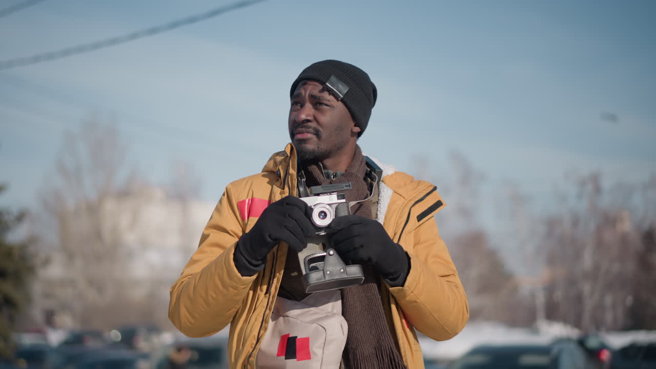 side view of black man in yellow jacket strolling urban street candidly scanning environment with vintage camera hanging by strap under clear winter sky beside parked cars and bare trees