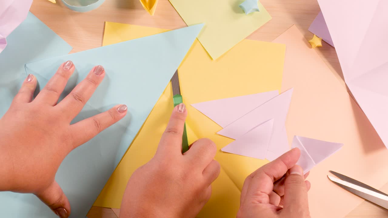 Two hands fold colored paper into origami shapes on a pastel desk under soft lighting