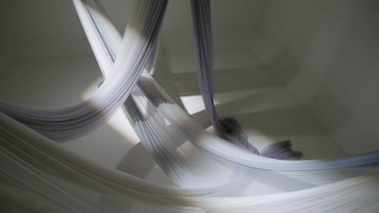 Ampia vista interna di un'installazione di tessuto plissettato sospeso con pannelli semitrasparenti drappeggiati dai punti del soffitto in anelli incrociati, pieghe raccolte, texture stratificata, ombre morbide e toni neutri e calmi.