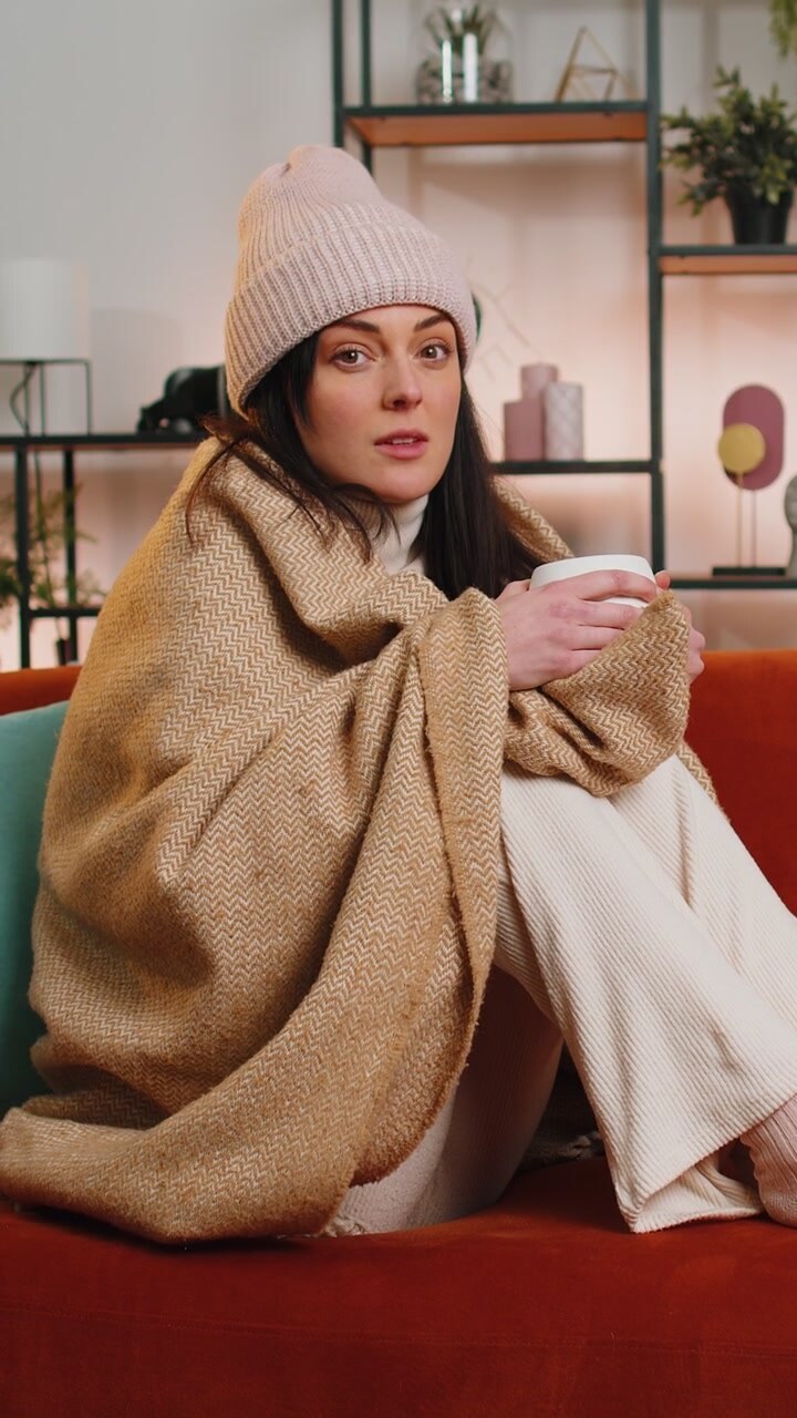 Una donna malata e malata indossa un cappello avvolto in un quadri si siede tremando di freddo sul divano a bere tè caldo
