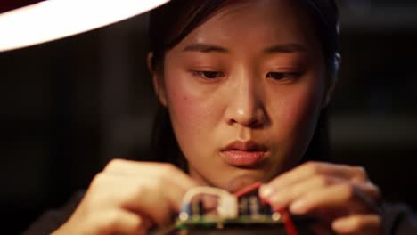 in a dimly lit workshop, a young woman concentrates on repairing a circuit board. she carefully connects wires while deep in thought, showcasing her technical skills and dedication to the craft.
