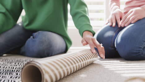 low section of biracial mother and adult daughter wrapping christmas gift at home, slow motion