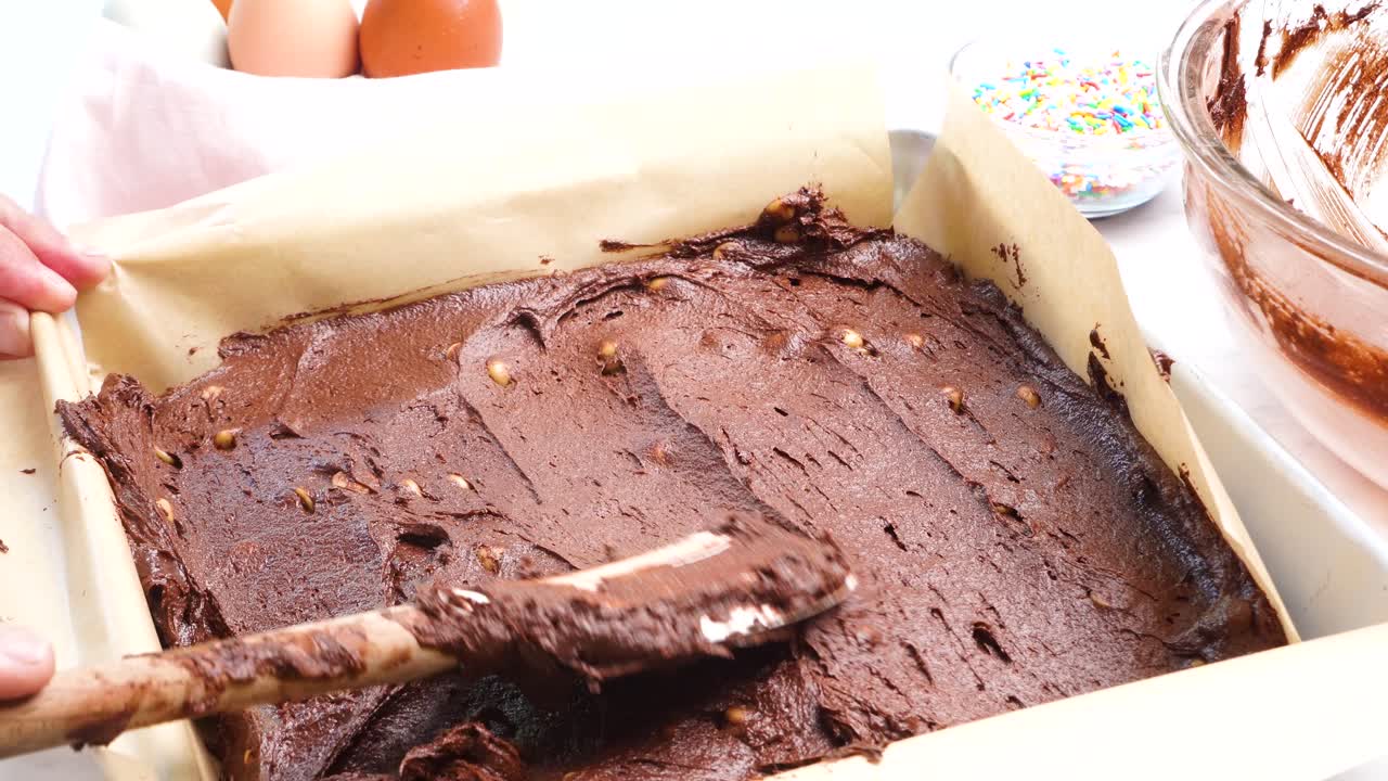 Baking chocolate brownies and using a spatula to spread out the batter. Making brownies on a white background and batter in the square baking pan. In the kitchen making a chocolate dessert