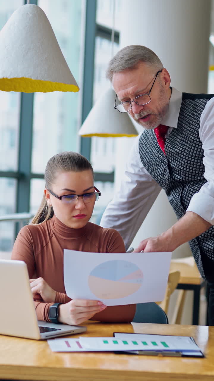 Young woman sitting at table and listening to her boss. Mature businessman with his leg on desk is talking to a secretary in the modern office. Vertical video