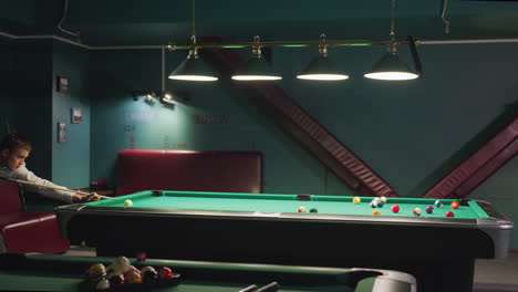 player in white shirt and grey trousers leans at edge of green pool table, gripping cue stick with intense focus. dark background enhances concentration as he strikes cue ball, aiming for precise