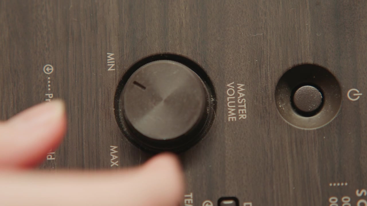 Human fingers rotate textured round control knob on dark wood-grain device panel beside recessed power button, with close framing, tactile grip, and subtle motion blur showing manual adjustment