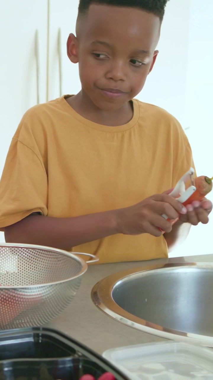Bambino che esamina un pelapatate, poi pela le carote producendo lunghe strisce, mentre un adulto prepara i pasti.