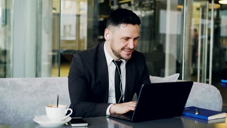 happy caucasian entrepreneur in formal clothes typing on his laptop while listening to music with earbuds in his ears in a modern cafe during lunch