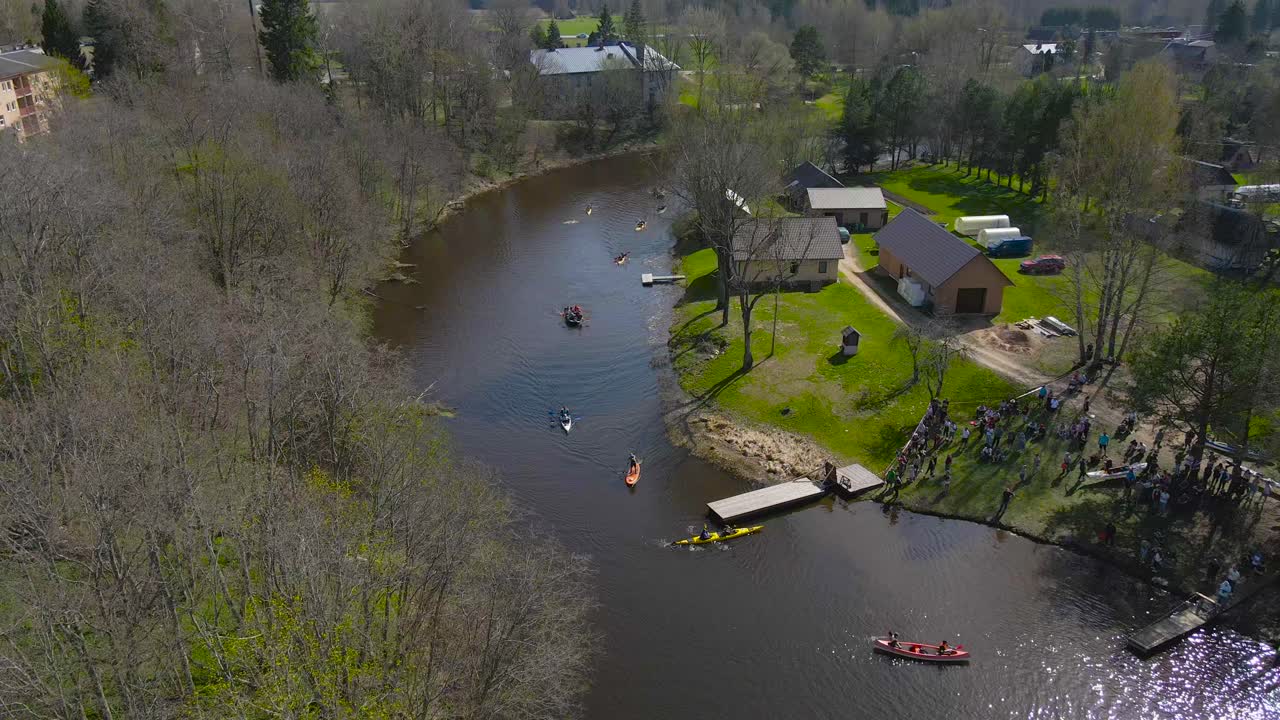 Gorgeous aerial drone view of colorful kayaks rowing and paddling to a wooden boat bridge during Võhandu marathon while the sun is shining on the narrow dark river. Green grass and houses visible.