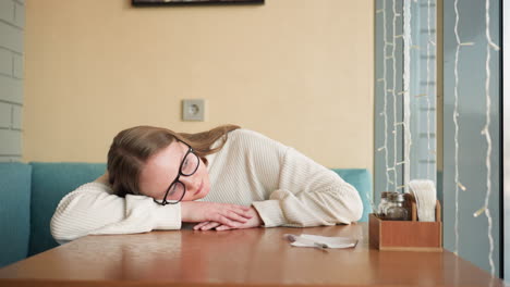 student wearing glasses and cozy white sweater resting head on wooden table inside brightly lit room near window with fairy lights, suggesting tiredness