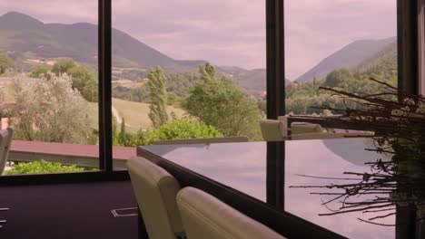 a wide shot of a modern dining room with a glass table and chairs, featuring a scenic view of provence’s rolling hills and forests through large windows at dusk.