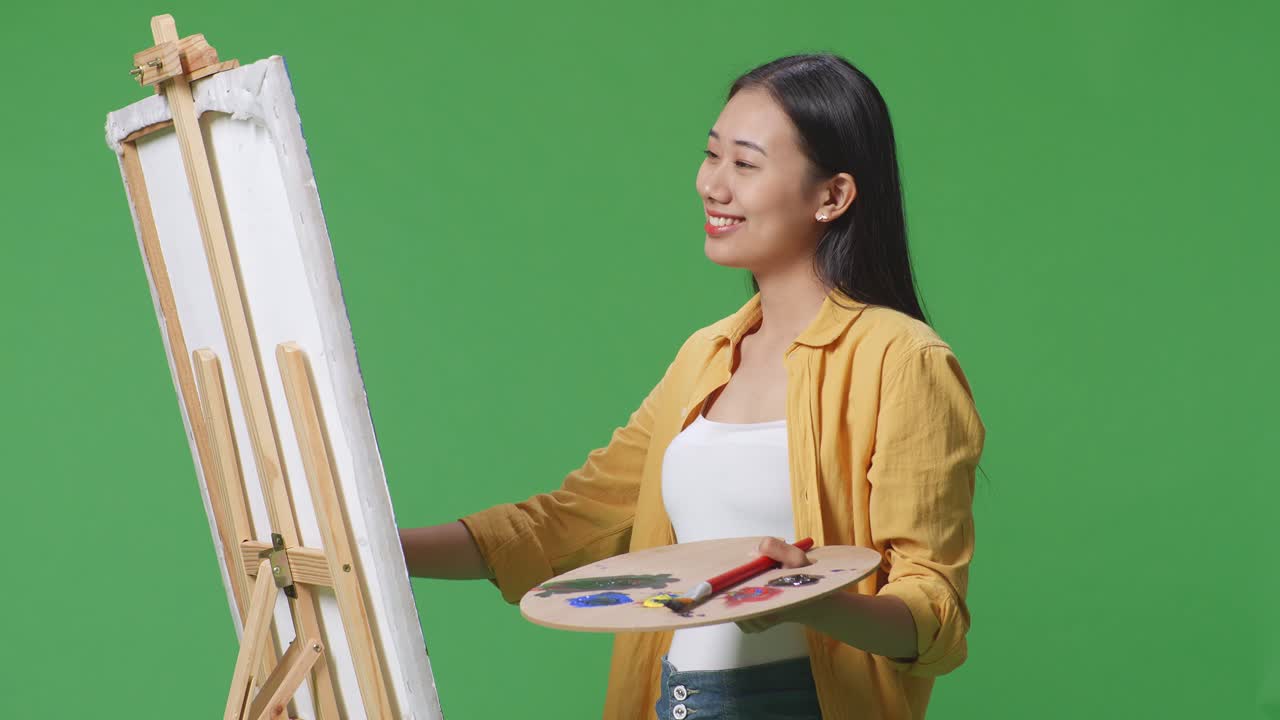 아시아 여성 예술가 가을 올리고 미소 짓고 캔버스에 기름색 페인트와 브러쉬를 그리며 그린 스크린 스튜디오에 있습니다.