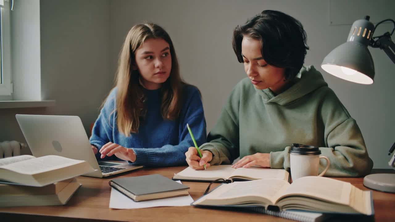 En person i blå genser skriver på laptop til venstre, og en i grønn hettegenser skriver med grønn blyant til høyre, snur seg for å avklare på hybelen.