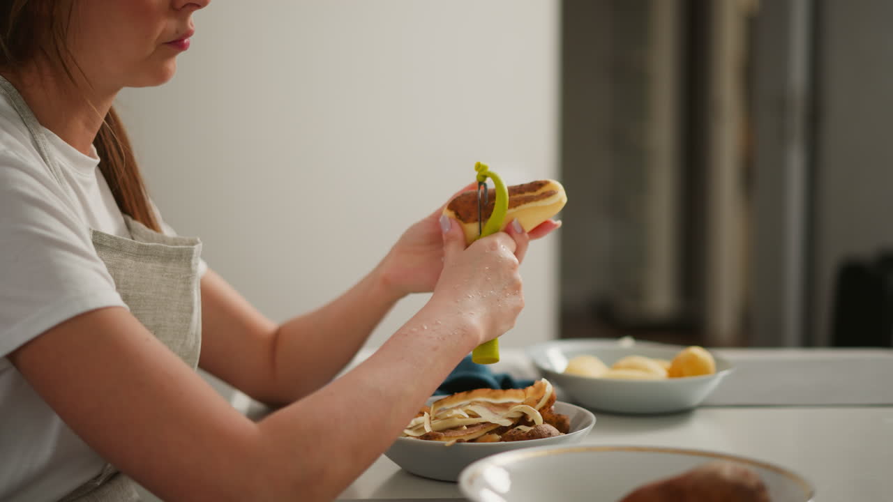 Portrait side view of woman in apron peeling potato over bowl filled with potato skins, soft blur background with open door, kitchen counter and other utensils partially visible