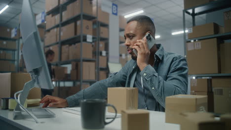 sorting center male african american warehouse associate talks to customer by mobile phone sorting center male african american warehouse associate talks to customer by mobile phone checks inventory on computer prepares package for delivery storage with orders of online ecommerce store