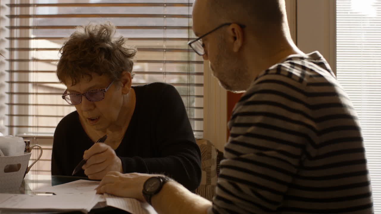 An elderly woman sits at a kitchen table with a middle-aged man. She signs documents with her left hand, as her caregiver son onlooking, steadies the page for her. Both wear reading glasses. 24fps.