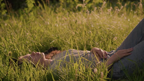 young woman lying in grassy field, relaxed with one hand resting on her leg and another on her stomach, surrounded by tall grass and bathed in warm sunlight