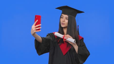 retrato de una estudiante en disfraz de graduación, sosteniendo un diploma y haciendo una selfie en el teléfono móvil. joven posando en el estudio con fondo de pantalla azul. primer plano. cámara lenta lista 59.94fps