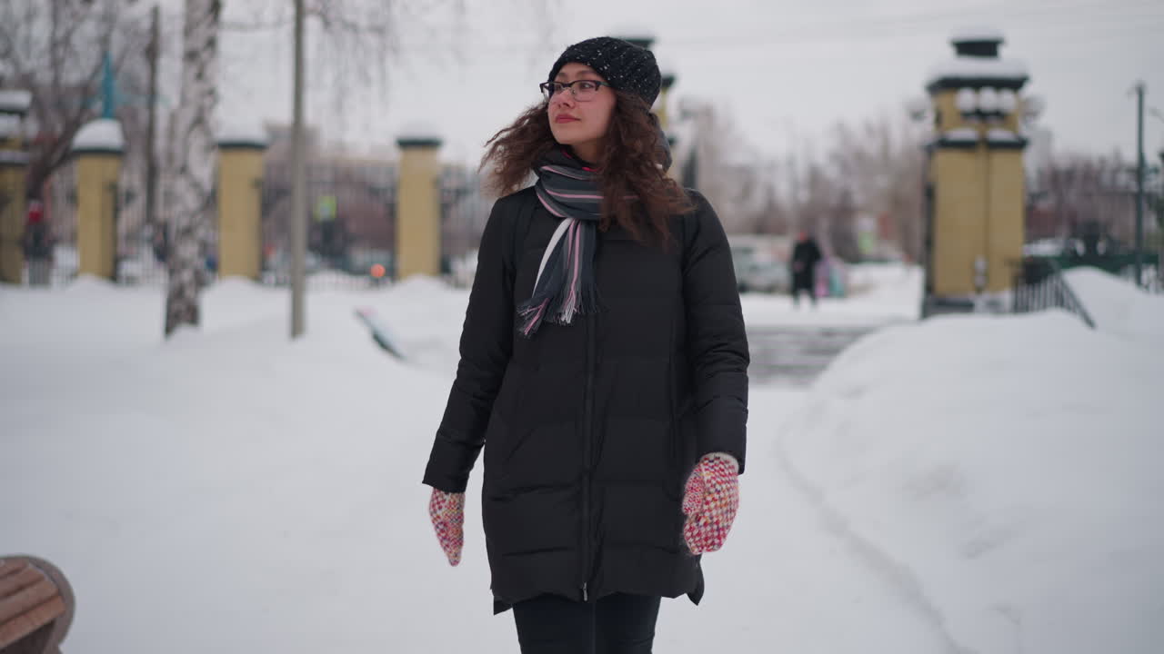 femme faisant signe de la main dans un parc enneigé en hiver, portant un manteau noir, une écharpe, un bonnet, des gants et des bottes, dans un environnement urbain, saison froide en extérieur, solitude, atmosphère glaciale, chemin gelé, environnement paisible, ciel nuageux