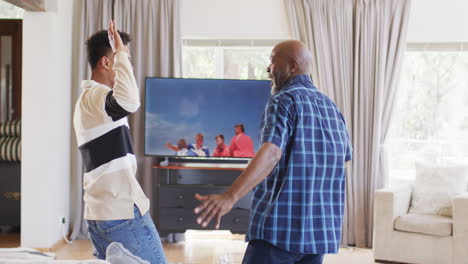 excited african american father and adult son cheering at sport on tv and high fiving, slow motion