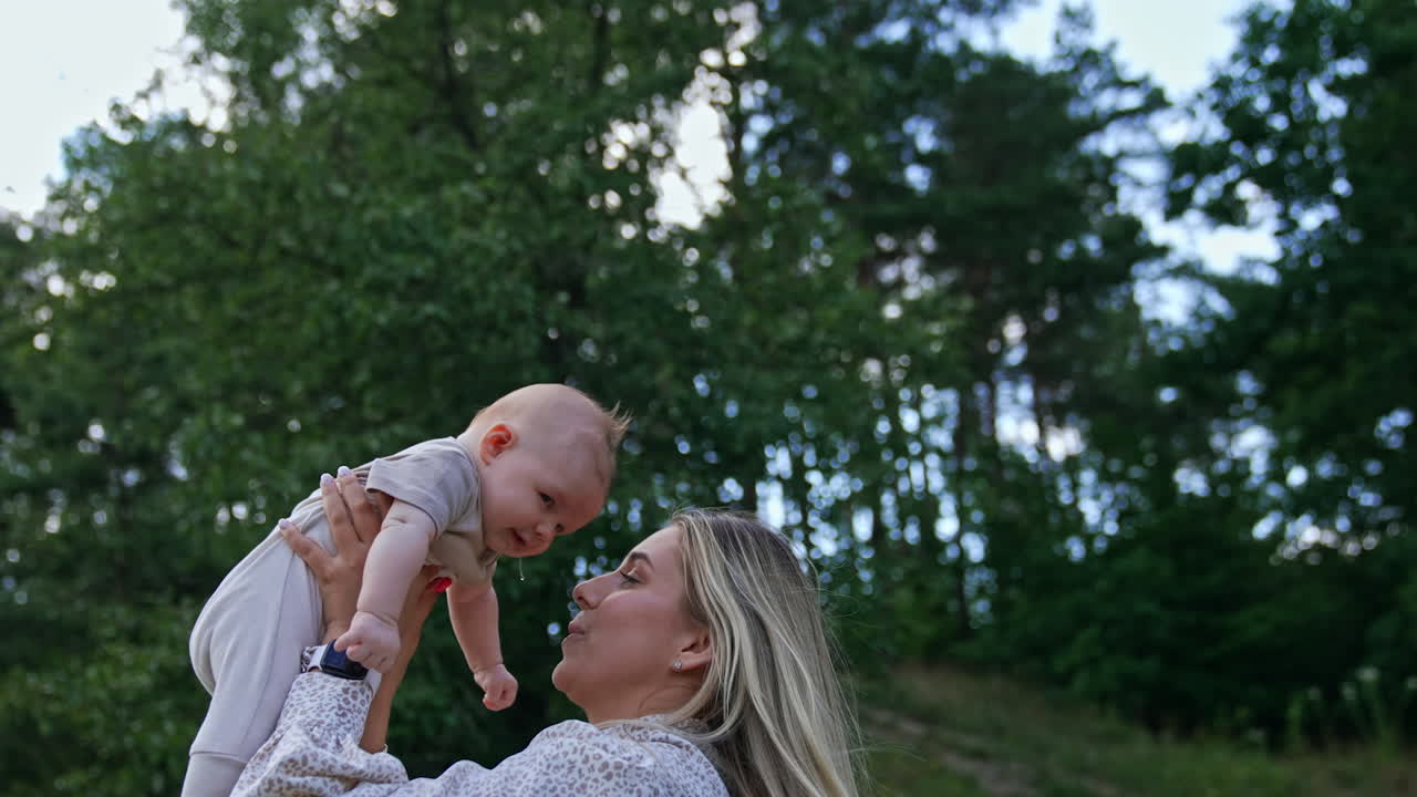 Blonde woman wearing light dress tossing her tiny kid. Happy woman playing with her child in nature.
