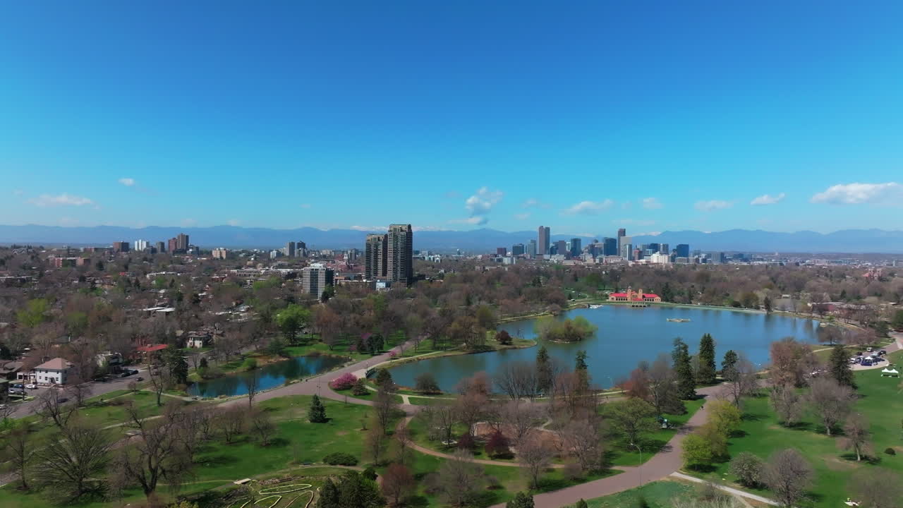 City Park Denver Colorado Lake Pavilion vibrant spring summer aerial drone sunny blue sky snow capped Rocky Mountains front range cityscape green lush grass trees blossom backwards motion