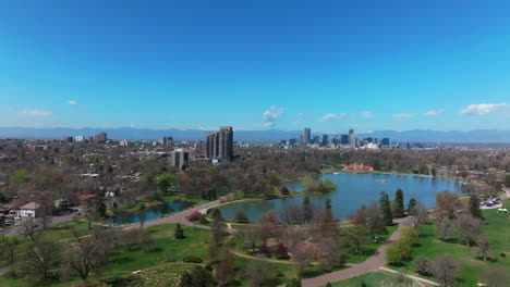 city park denver colorado lake pavilion vibrant spring summer aerial drone sunny blue sky snow capped rocky mountains front range cityscape green lush grass trees blossom backwards motion