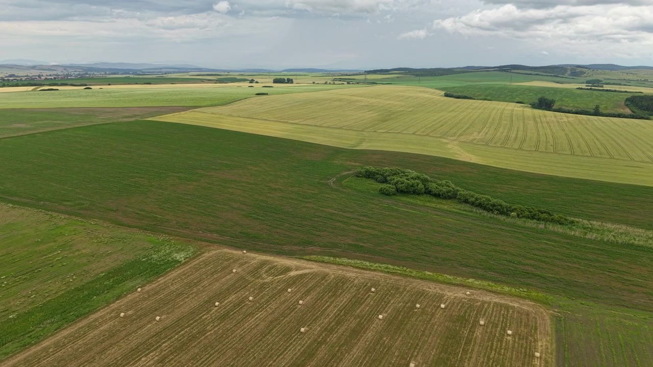 An aerial view reveals Hungary's diverse agricultural landscape, showcasing harvested fields, hay bales, and rolling green hills beneath a cloudy summer sky.