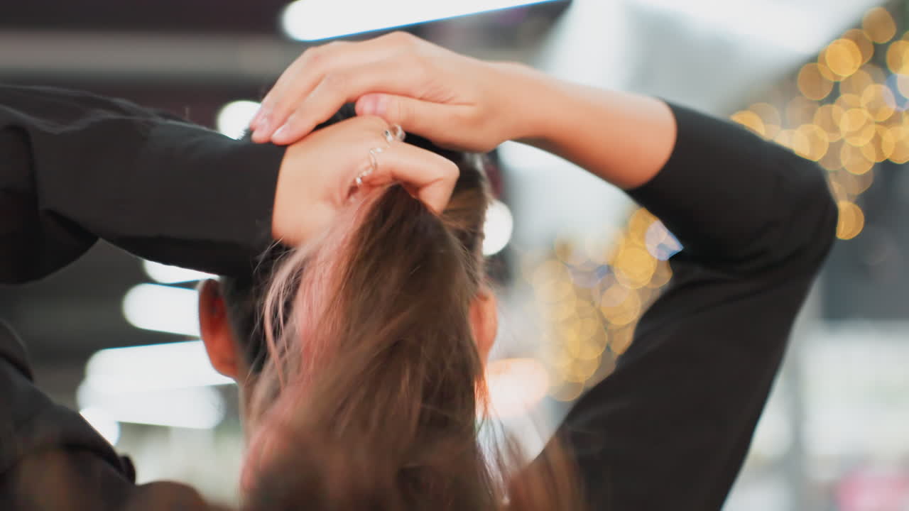 student tying ponytail in mirror, closeup back of head with hands gathering brunette hair, pink highlights peeking through, cozy indoor bokeh lights, casual sweater, preparation for campus routine