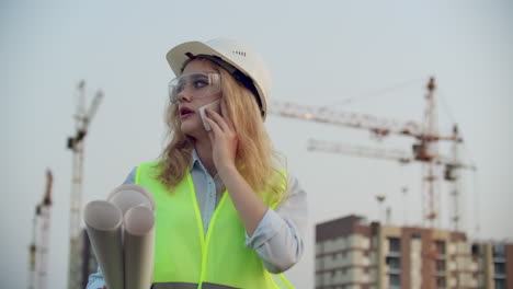 woman engineer designer talking on the phone with the contractor with drawings in hand on the background of buildings under construction and cranes