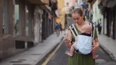 Carrying-her-infant-in-a-kangaroo-backpack,-a-young-woman-captures-memories-on-her-mobile-phone-during-her-travels.-As-she-walks,-she-glances-at-the-phone's-screen