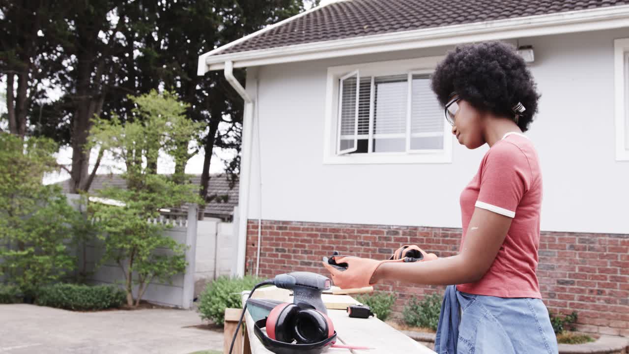 Mulher afro-americana examinando um banco em casa, pegando luvas e verificando uma ferramenta perto da lixadeira.