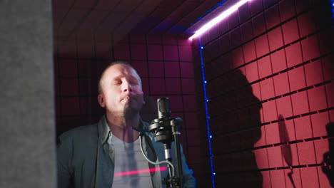 a man in a blue long-sleeve shirt and white inner shirt holds a microphone in a red soundproof studio, ready to sing. the dramatic lighting emphasizes the intense atmosphere of a recording session