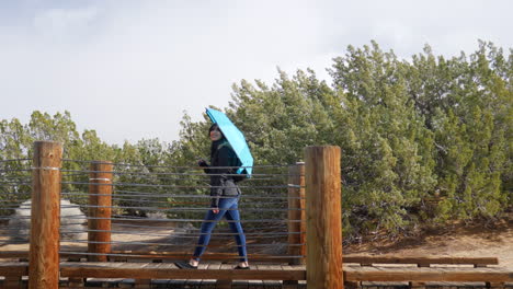 Eine-Schöne-Junge-Frau-Mit-Blauem-Regenschirm,-Die-Während-Eines-Regensturms-In-Der-Zeitlupe-Der-Wüste-über-Eine-Brücke-Geht