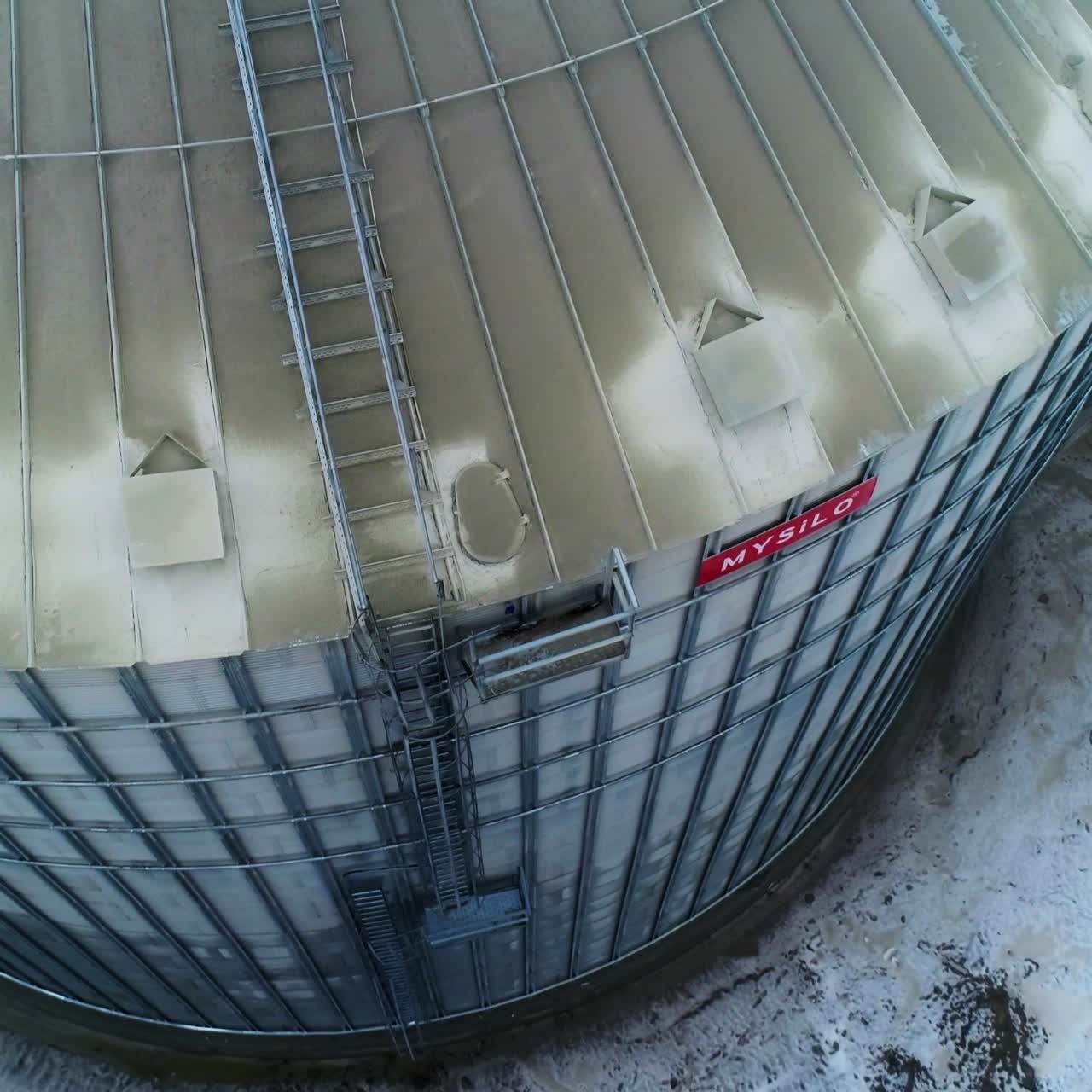 Flying around the enormous metal tank at agricultural elevator. Aerial footage over the dusty silo. Snowy ground backdrop