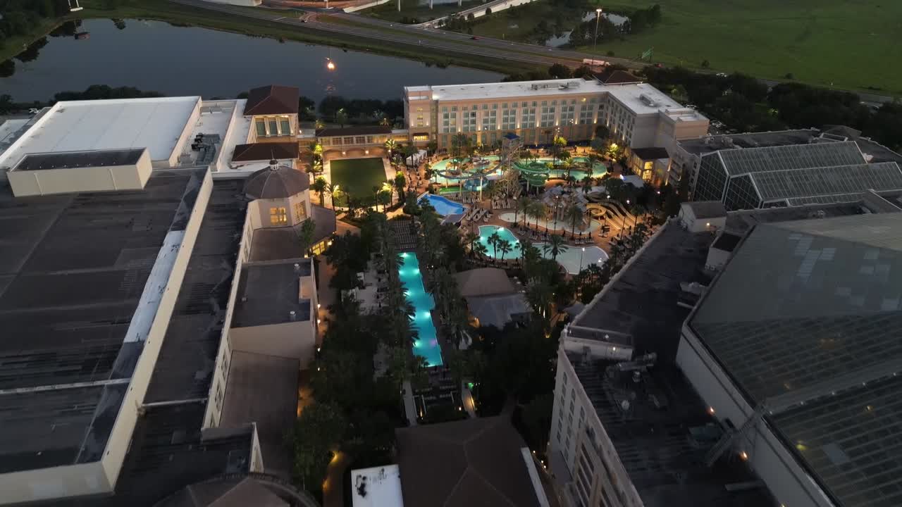 Aerial drone shot captures illuminated resort with pools, surrounding buildings, and nearby highway at dusk in Hawaii. Warm lighting contrasts with evening sky, creating a serene view.