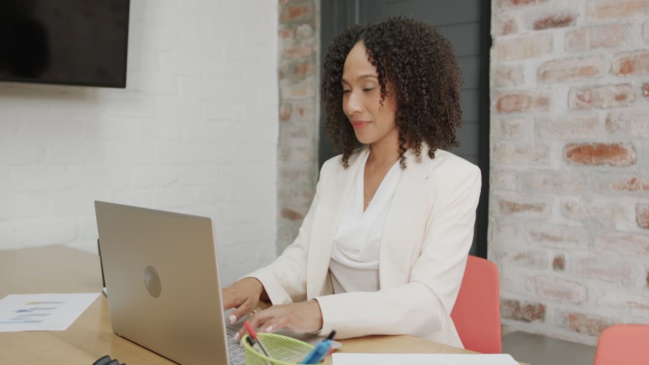 Retrato de empresária birracial à mesa usando um laptop e sorrindo no escritório, em câmera lenta