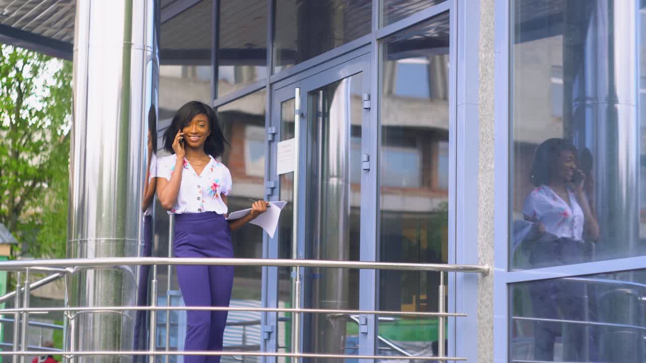 una giovane donna d'affari adulta afroamericana sta parlando al telefono fuori vicino a un moderno edificio per uffici.